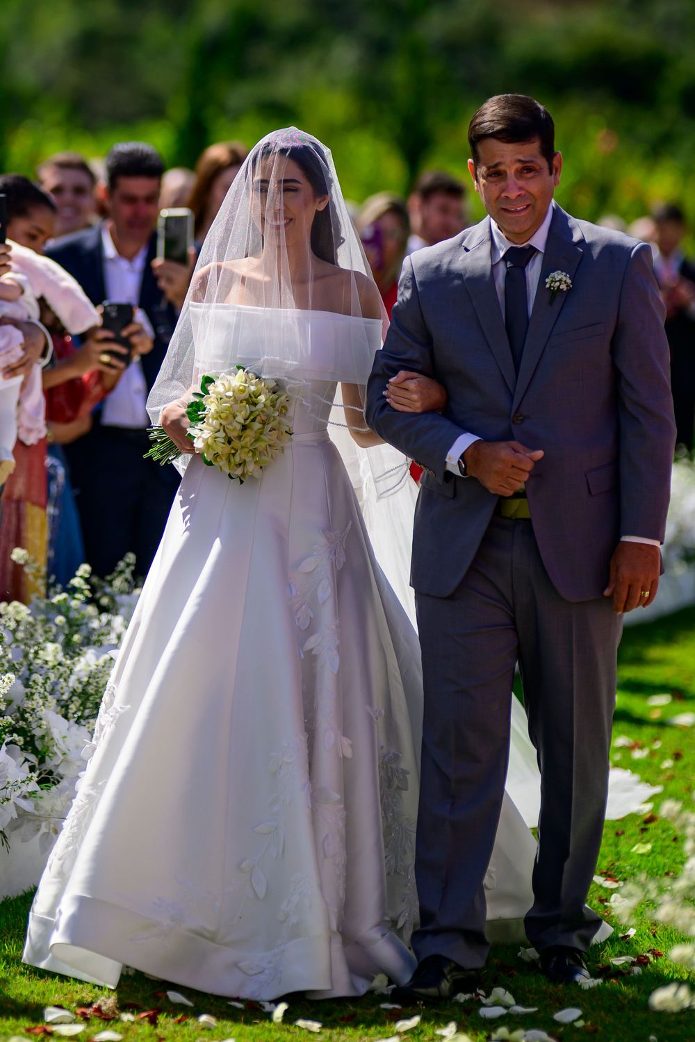 Elegant bride in off-shoulder gown with veil walks down outdoor aisle with father.
