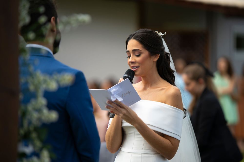 Beautiful bride in off-shoulder wedding dress reads heartfelt vows at outdoor ceremony.