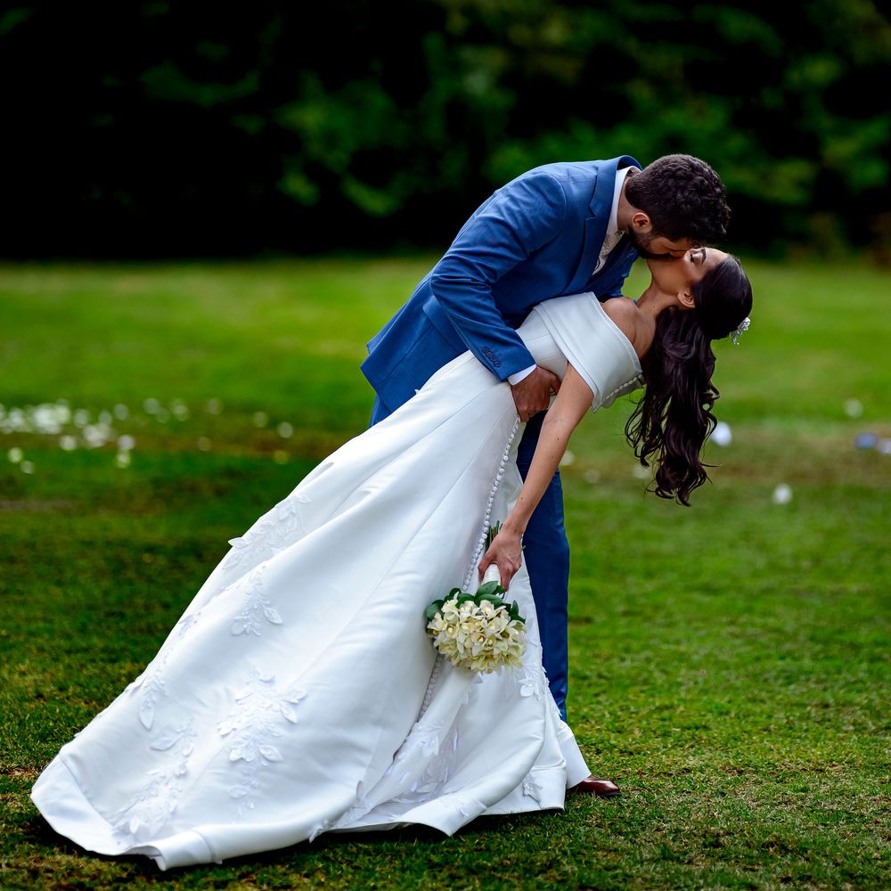 Groom in blue suit dips bride in white gown for romantic kiss on green lawn.