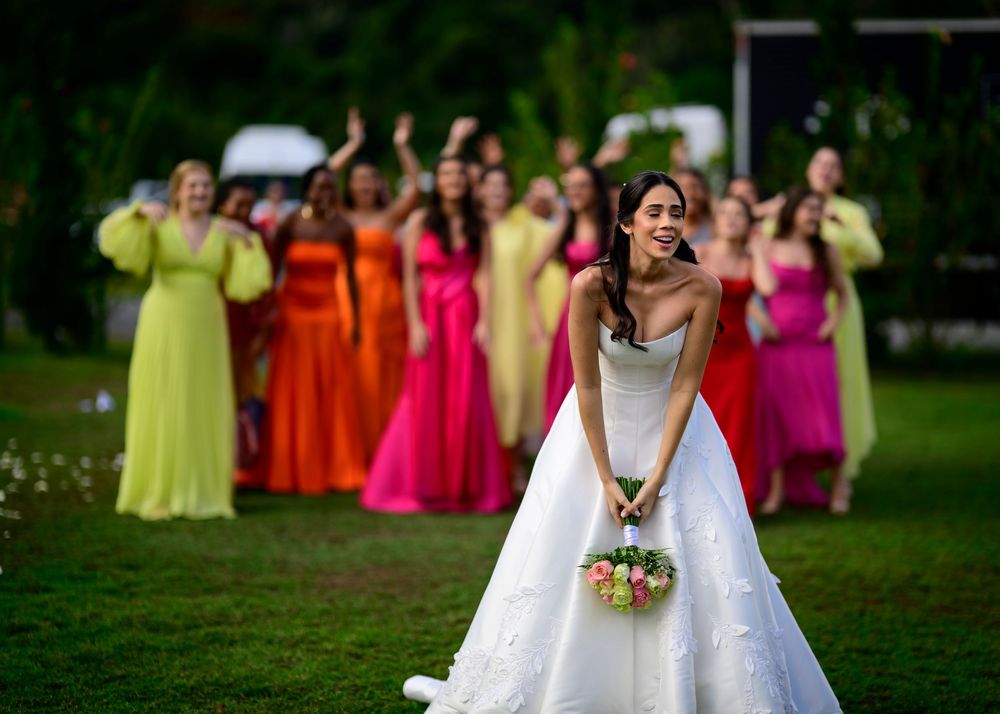 Smiling bride in white gown tosses bouquet to colorfully dressed bridesmaids outdoors.
