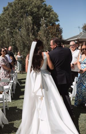 Wedding ceremony guests stand in rows of white chairs on a sunny lawn surrounded by trees.