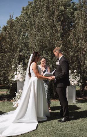 An outdoor wedding ceremony takes place in a garden setting with trees and decorative statues in the background.