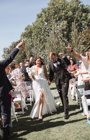 Wedding guests celebrate by throwing confetti on a sunny day at an outdoor ceremony on green grass.