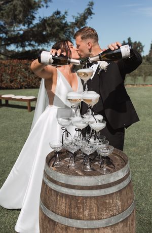 A couple in wedding attire pours champagne into a tower of glasses arranged on a rustic wooden barrel outdoors.