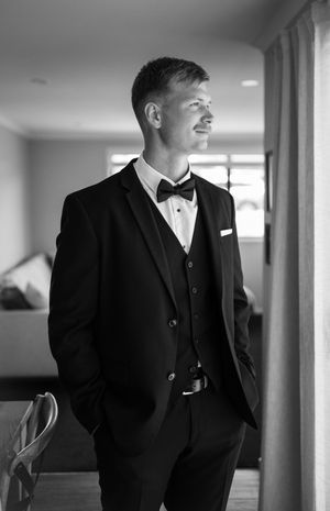 Black and white photo series of a groom in a tuxedo standing by a window before his wedding ceremony.