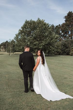 A couple in wedding attire holds hands while walking on a grassy lawn with trees in the background.