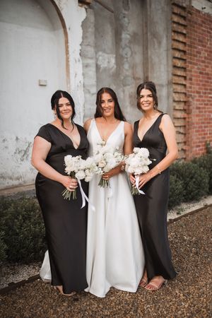 Wedding party members in black dresses and white gown hold bouquets against a brick and stone wall backdrop.