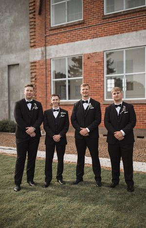 Groomsmen in black tuxedos line up for a formal photo against a brick building exterior.