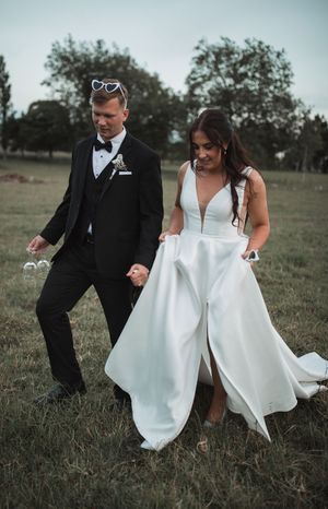 Newlyweds walking together on grass, bride in flowing white dress and groom in classic black tuxedo.
