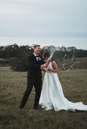 Newlyweds celebrate with champagne spray in an outdoor field during golden hour.