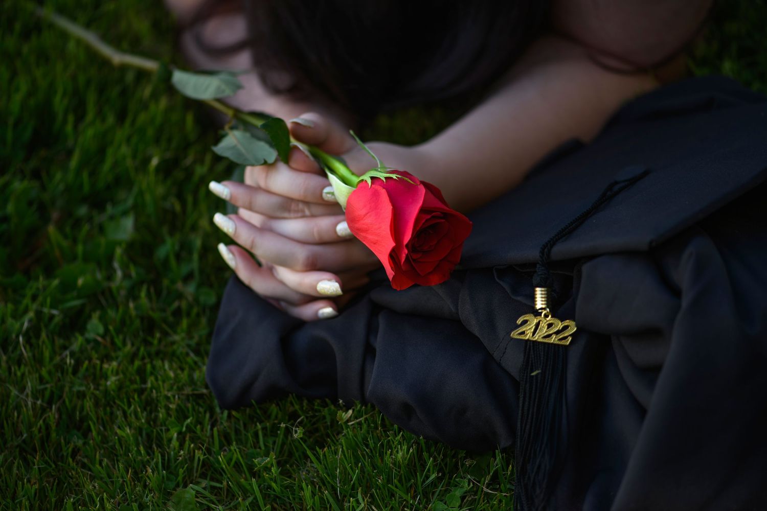 Graduate in black gown holding a red rose while sitting on green grass outdoors.