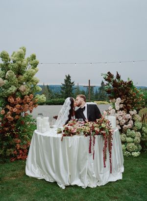 White tablescapes adorned with lush floral arrangements and string lights create a romantic outdoor wedding reception setting.