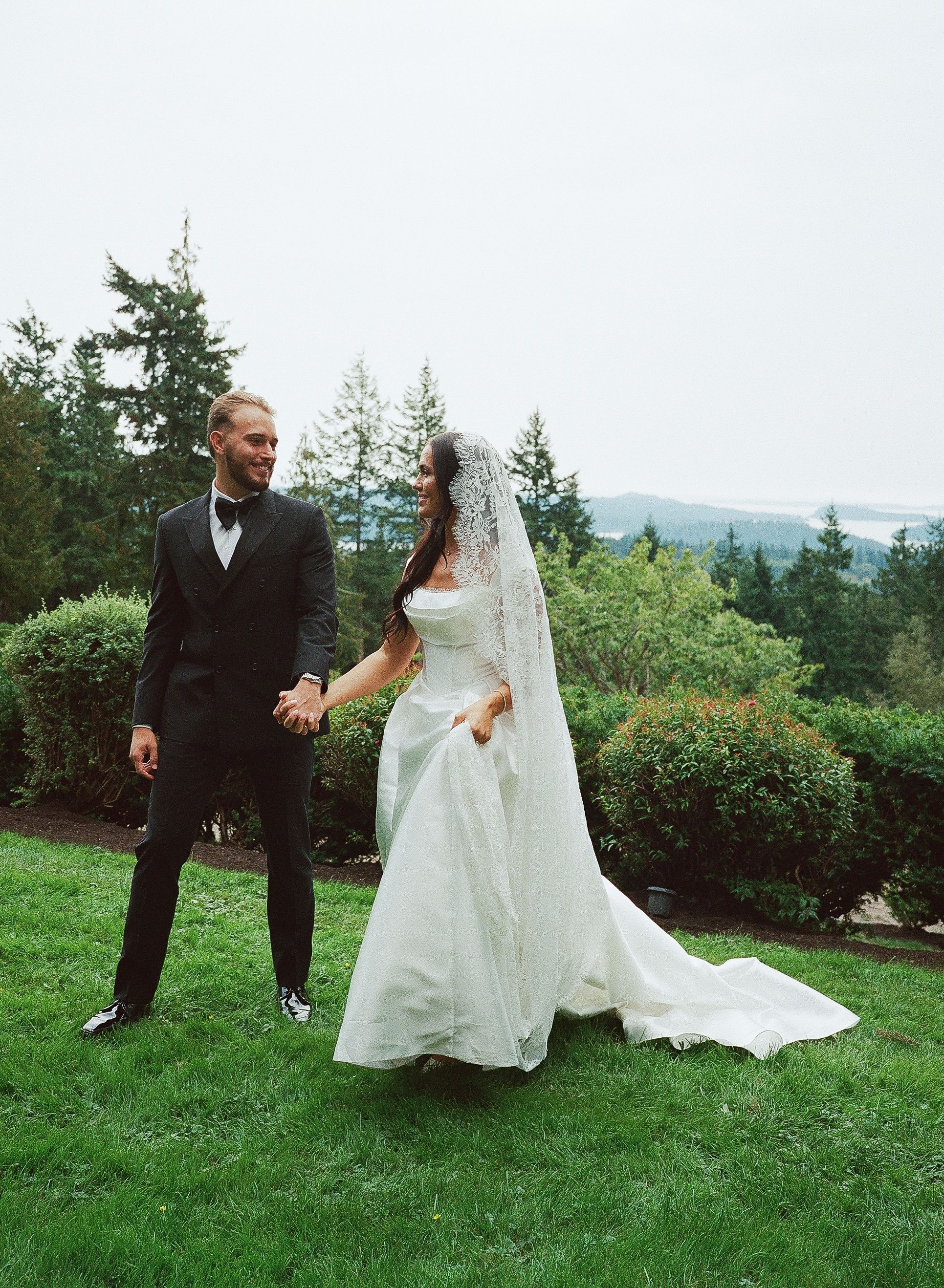 Newlyweds dance and twirl on a grassy hilltop with evergreen trees in the background.