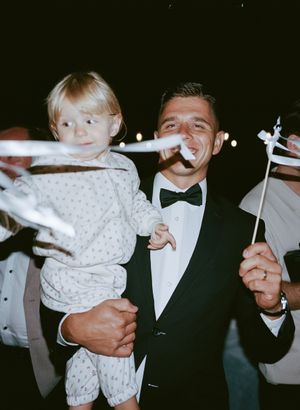 Wedding reception guests in formal attire hold and play with a small child in white clothing.
