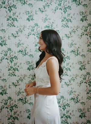 A bride in a white lace dress poses against a floral wallpaper background.