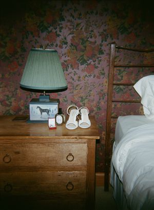 A vintage wooden nightstand with a mint green table lamp sits beside a white bed against floral wallpaper.