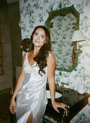 Woman in white silk nightgown poses playfully against floral wallpaper background.