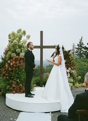 A wedding ceremony takes place on an elevated platform with a large wooden cross and floral arrangements.