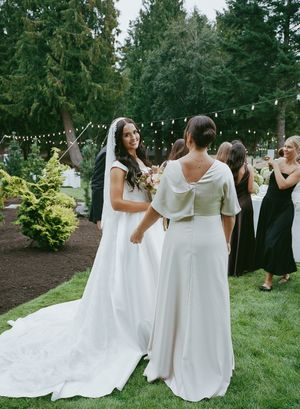 Bridesmaids in white dresses walking together on grassy lawn with evergreen trees in background.