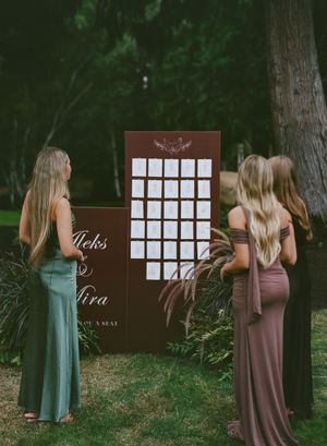 Wedding seating chart display with wooden boards in a forest setting surrounded by bridesmaids in pastel dresses.