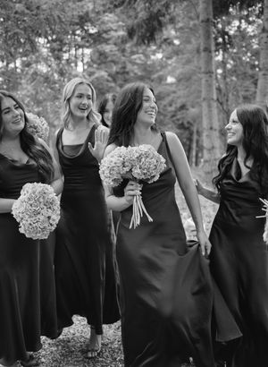 Bridesmaids in black dresses holding white hydrangea bouquets pose together outdoors.