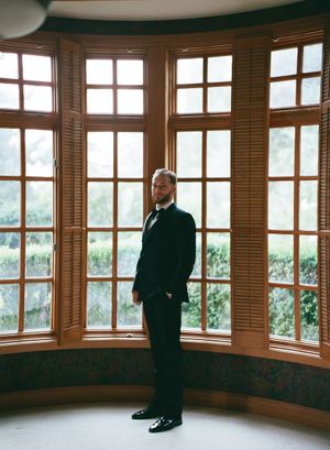 A person in a dark suit stands in a corner bay window area with wooden trim and natural light streaming in.