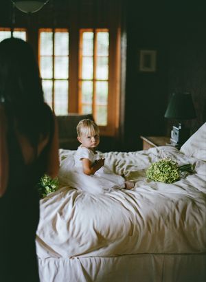 Small child in white dress sits on unmade bed with green floral arrangements in moody interior lighting.