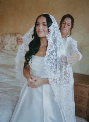 A delicate white lace veil flows elegantly around two figures in white dresses creating an ethereal bridal moment.