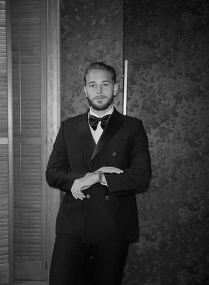 Four men in black suits and bow ties pose in a formal portrait against a textured wall.