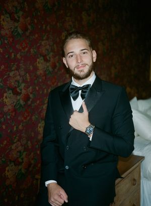 A groom in a classic black tuxedo and bow tie poses against a dark floral wallpaper background.