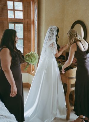 Wedding party members in black dresses help adjust a flowing white wedding gown in a sunlit room.