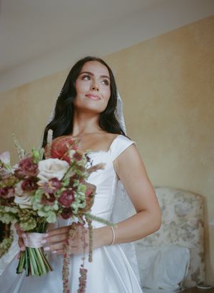 A white bridal bouquet with pink and burgundy flowers is held against a light colored background.