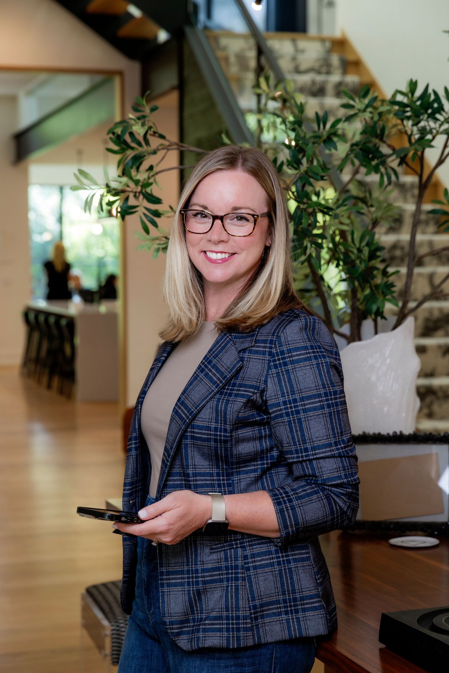 Professional wearing a plaid blazer and glasses sits at a desk in a modern office space with natural lighting.