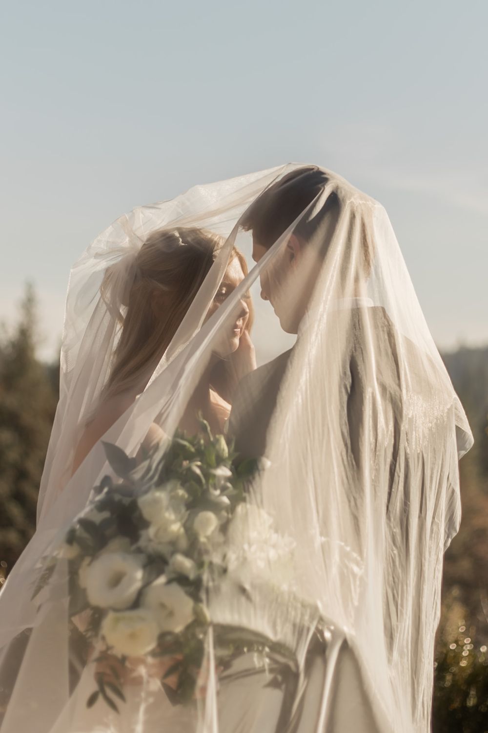 A romantic wedding portrait showing a couple sharing an intimate moment under a flowing white veil with soft sunlight.