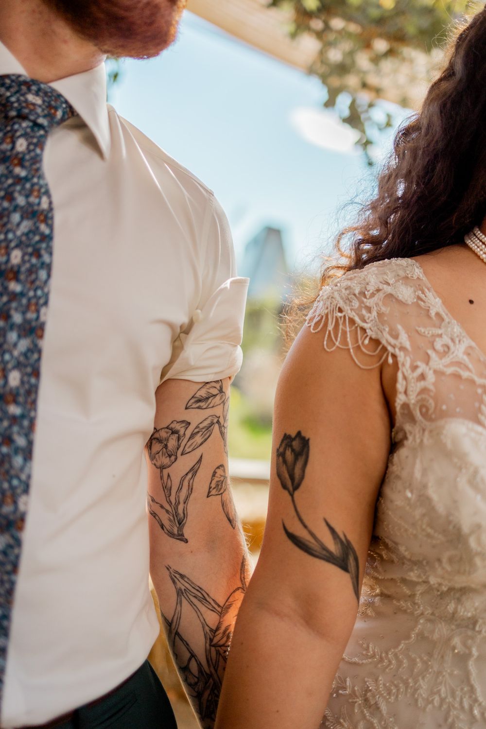 Close-up view of tattooed arms and wedding attire details showing lace dress and white shirt at an outdoor ceremony.
