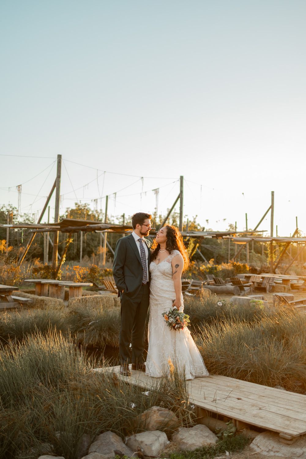 A couple in wedding attire stands on a rustic wooden walkway at sunset in a desert landscape setting.