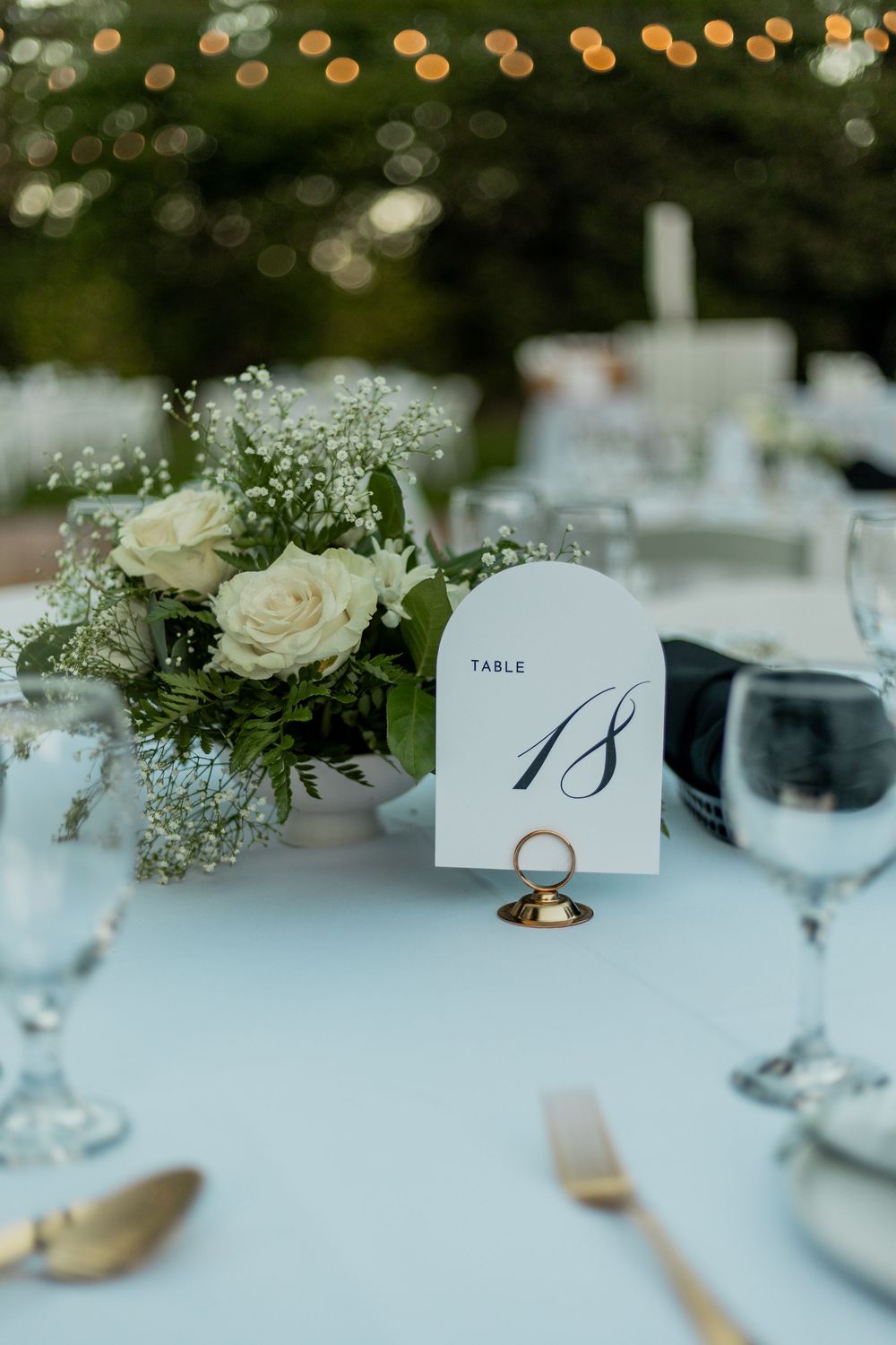 Elegant wedding table setting with white roses centerpiece, table number 18, and crystal glassware on white tablecloth.