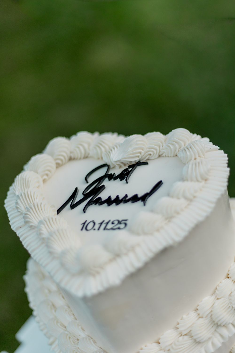 A white heart-shaped cake with decorative piped frosting and black writing on top against a green background.