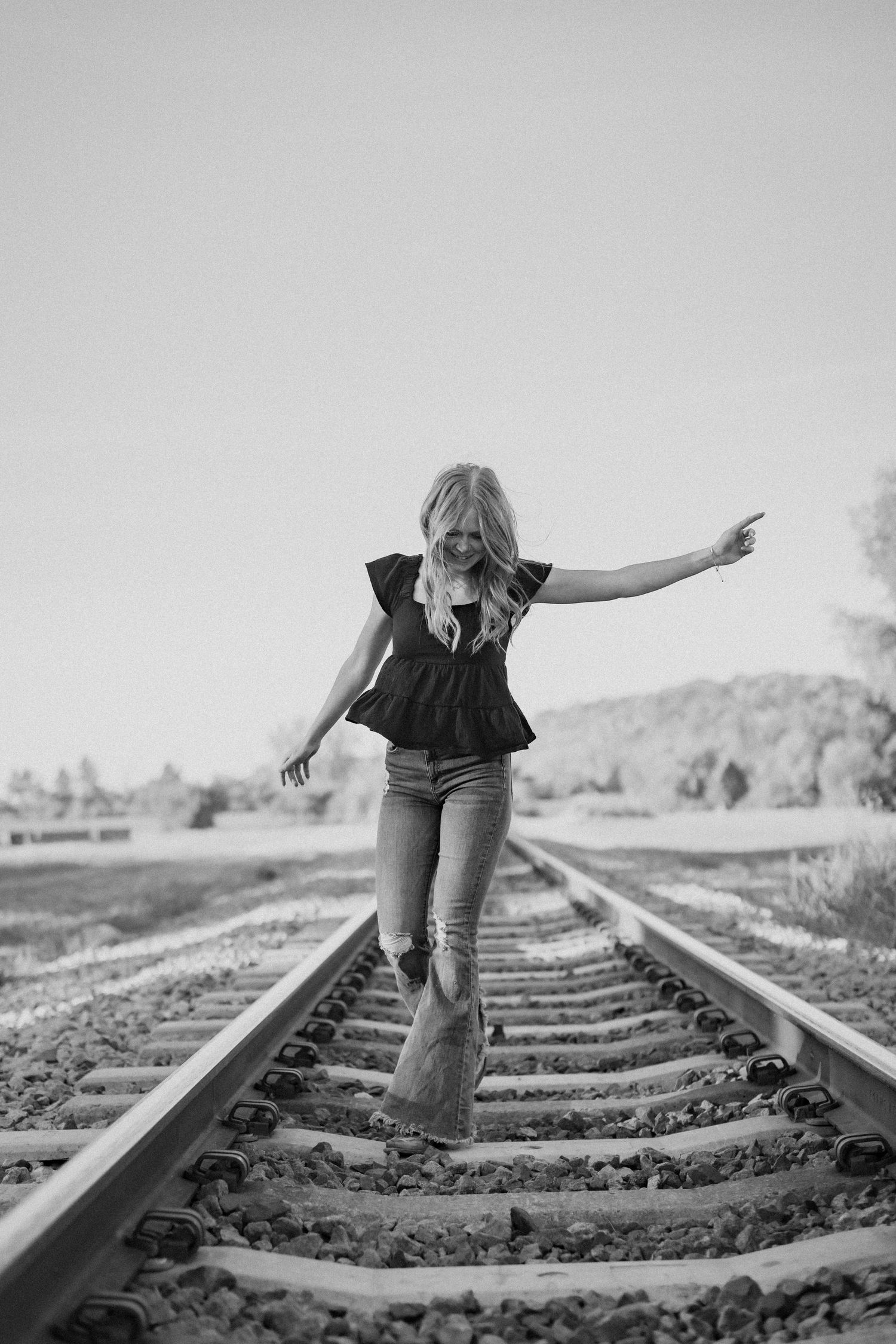 A young woman with arms outstretched walks along the railroad tracks in this black-and-white edit from her senior photos in Kansas City, Mo.