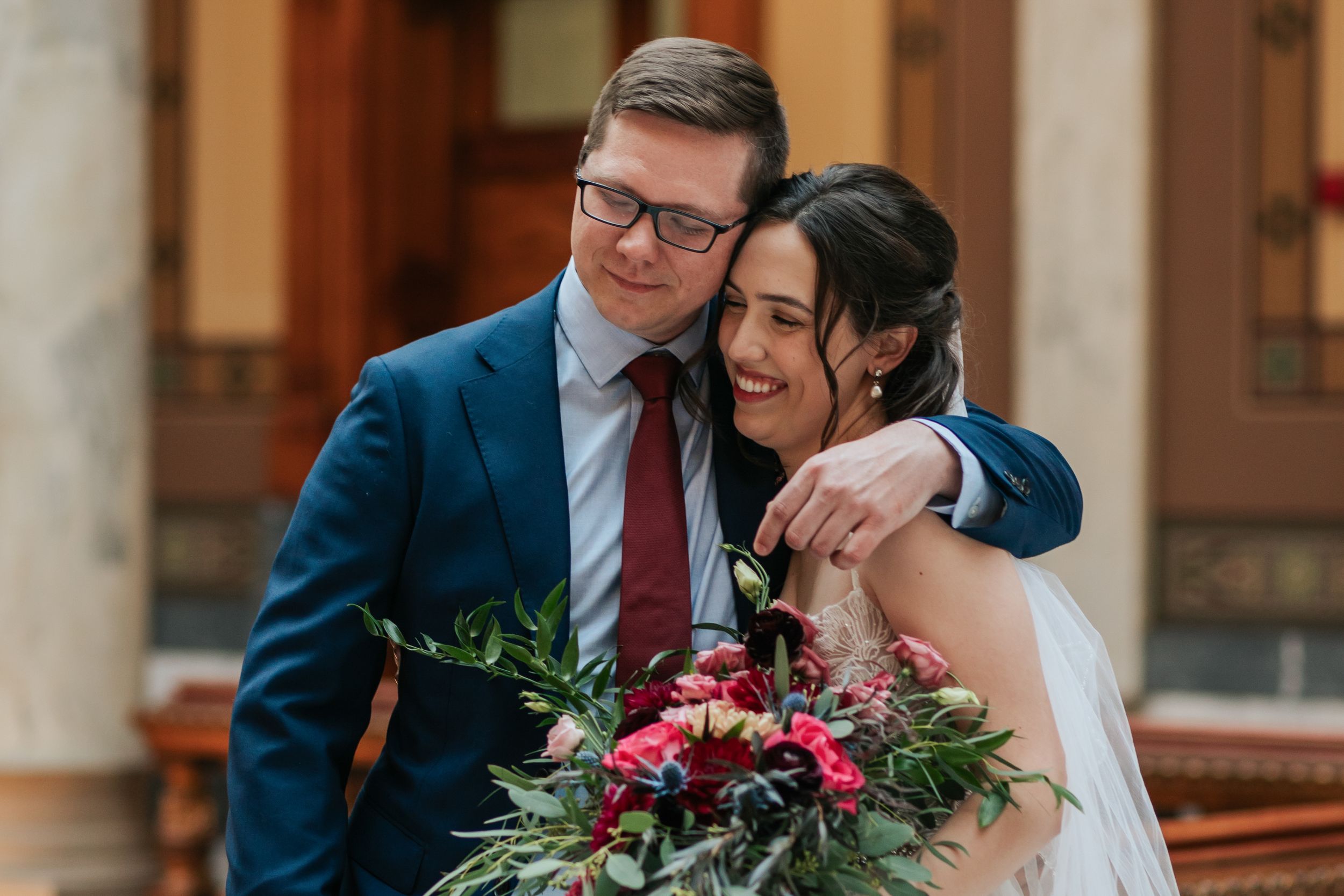 A happy couple embraces at their wedding, with the bride holding a vibrant red and green floral bouquet.