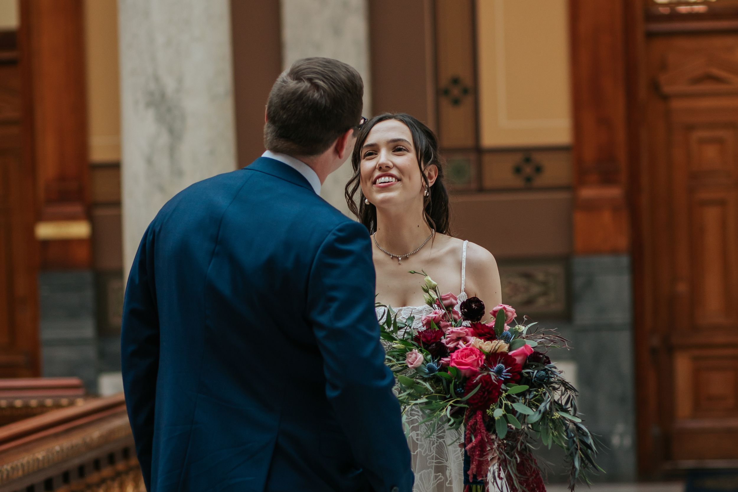 A bride and groom share an intimate moment while holding a vibrant red bouquet in an elegant historic building.