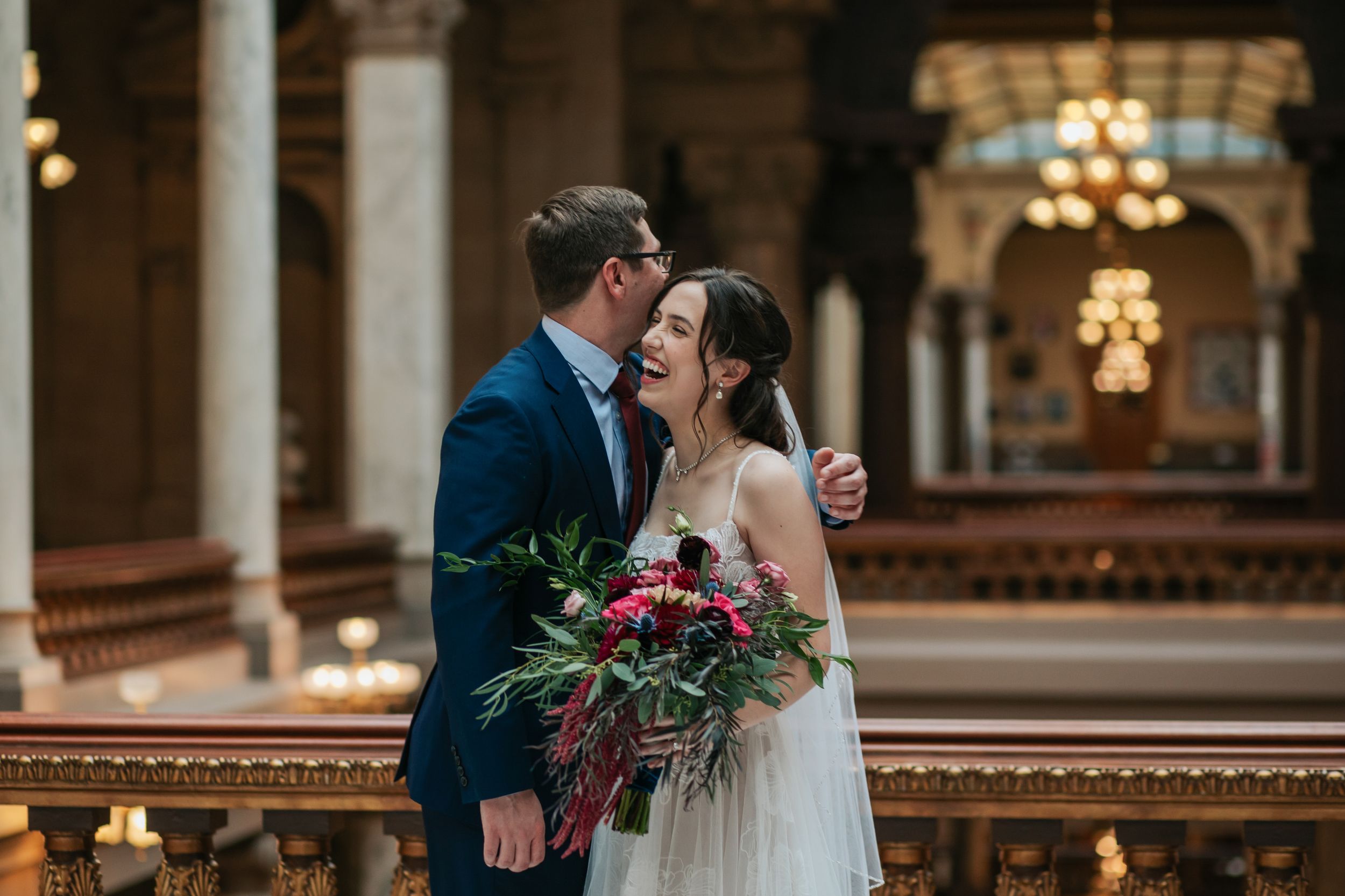 Newlyweds embrace in a grand historic hall with beautiful columns and chandeliers overhead.