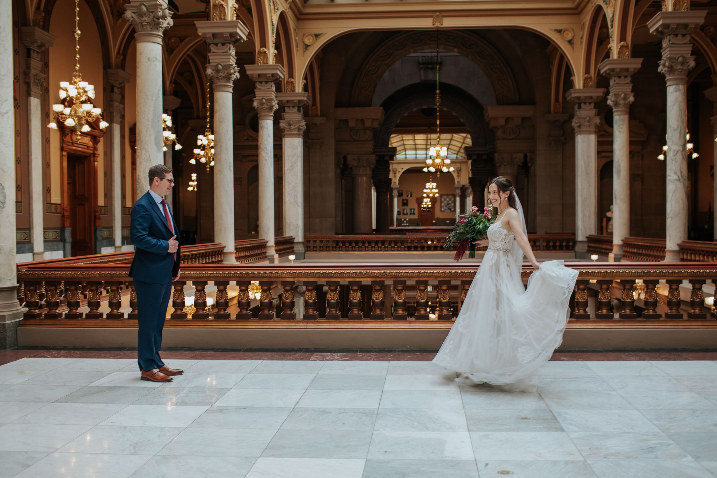 Elegant wedding couple poses in ornate historic building with marble columns and grand chandeliers.