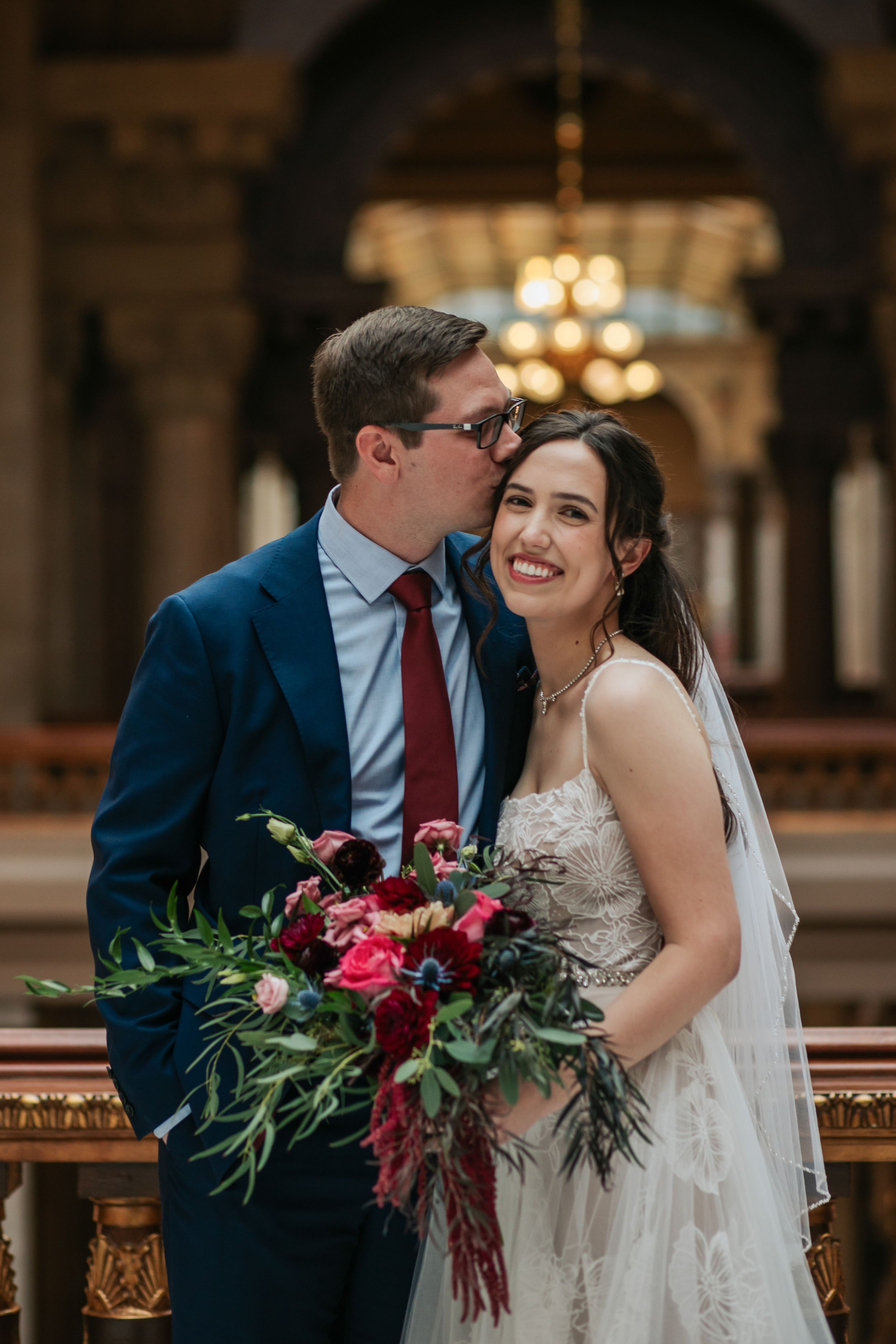 A romantic wedding couple sharing a tender moment in an ornate venue with a burgundy and pink floral bouquet.