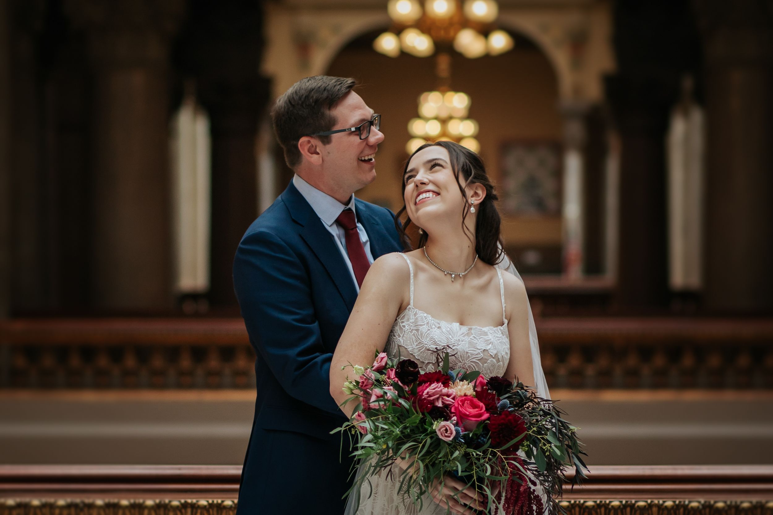 Couple in wedding attire shares romantic moment in front of elegant building with ornate chandeliers.