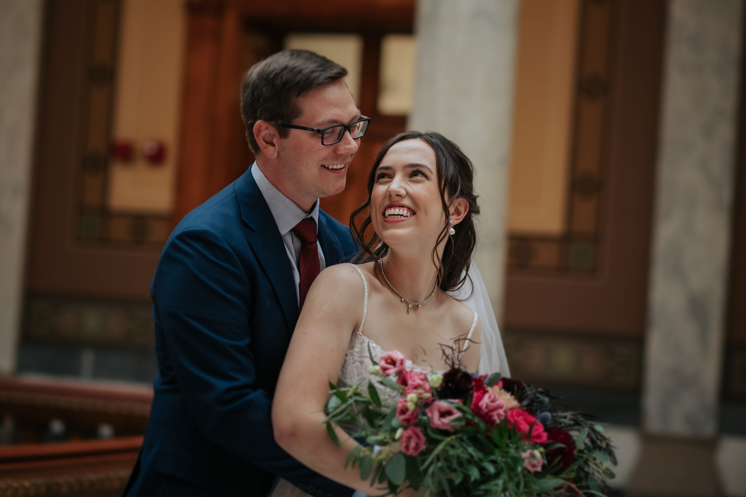 Happy couple laughs together during their wedding photoshoot in an ornate indoor setting.