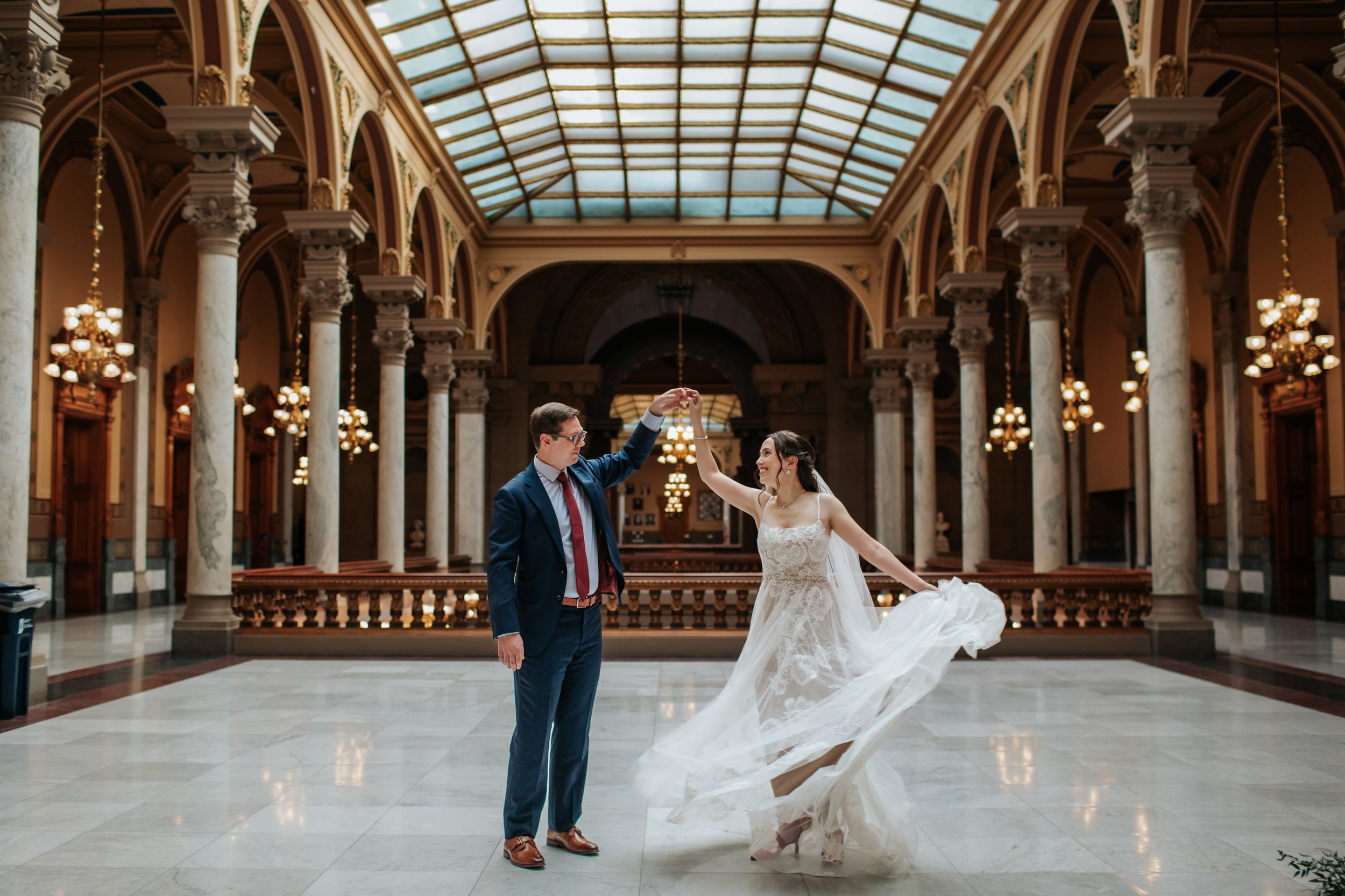 Newlyweds pose in a grand historic hall with glass ceiling and elegant architectural details.