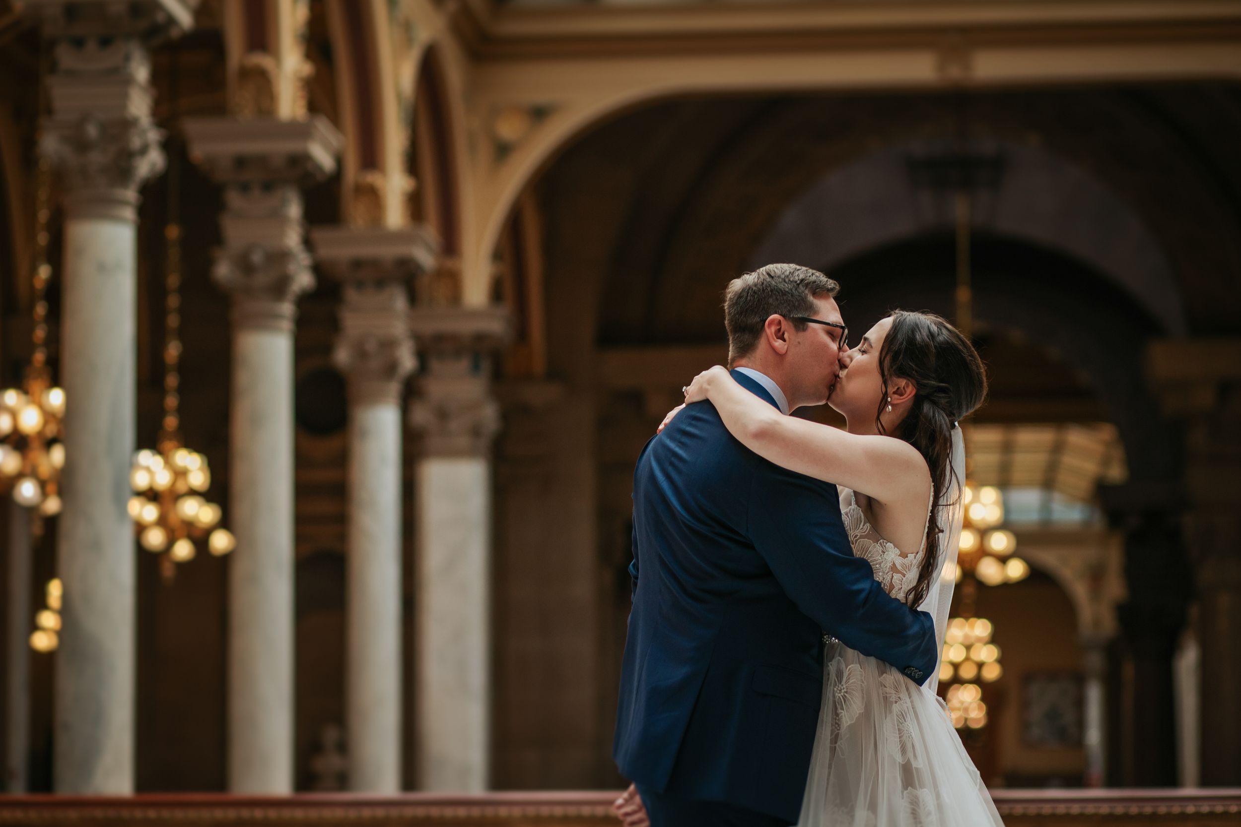A couple shares their first dance in an elegant ballroom with ornate columns and chandeliers.