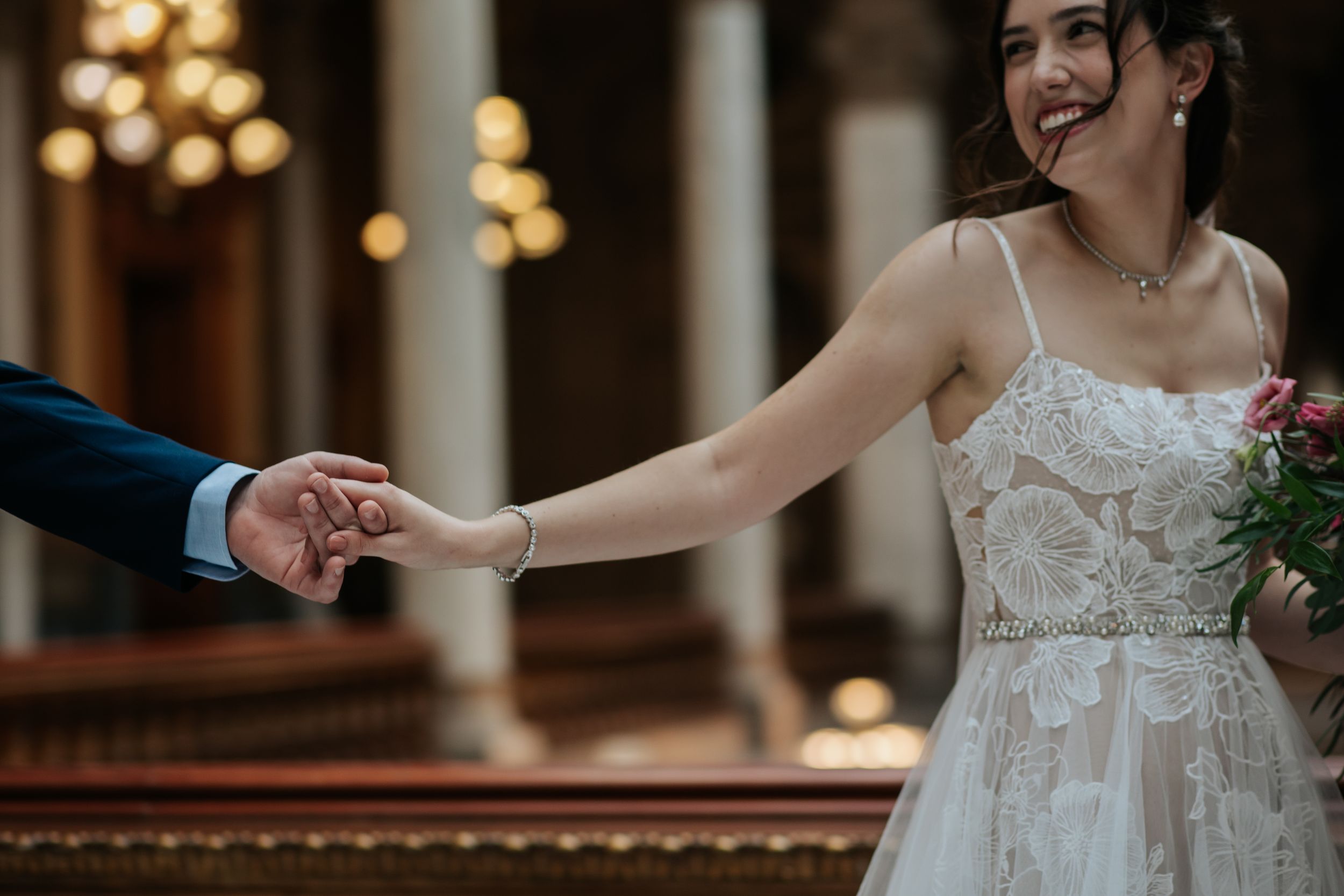 A bride and groom dance together in an ornate ballroom with classical architecture and warm lighting.