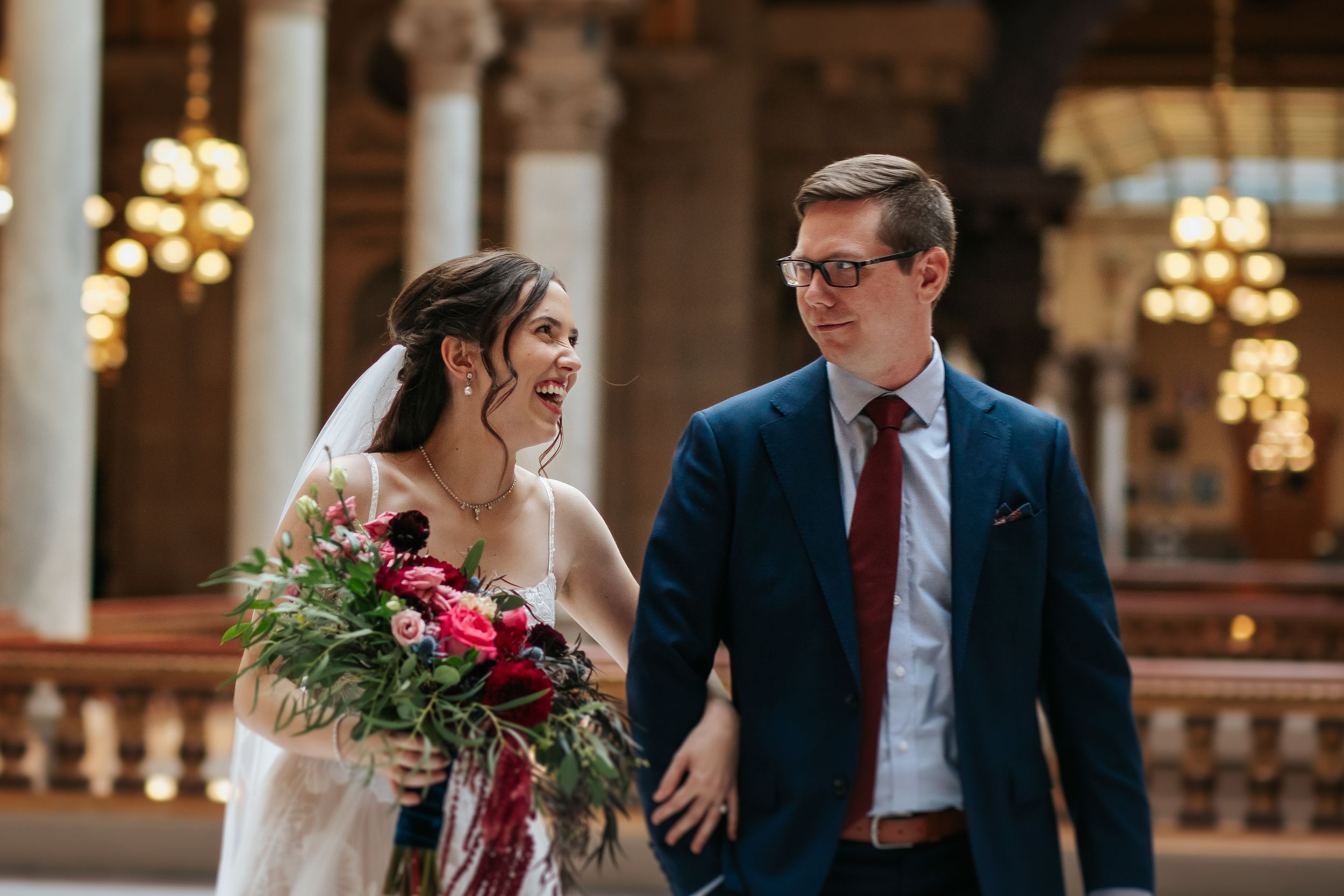 Bride with bouquet and groom share an intimate moment in a grand hall with chandeliers.